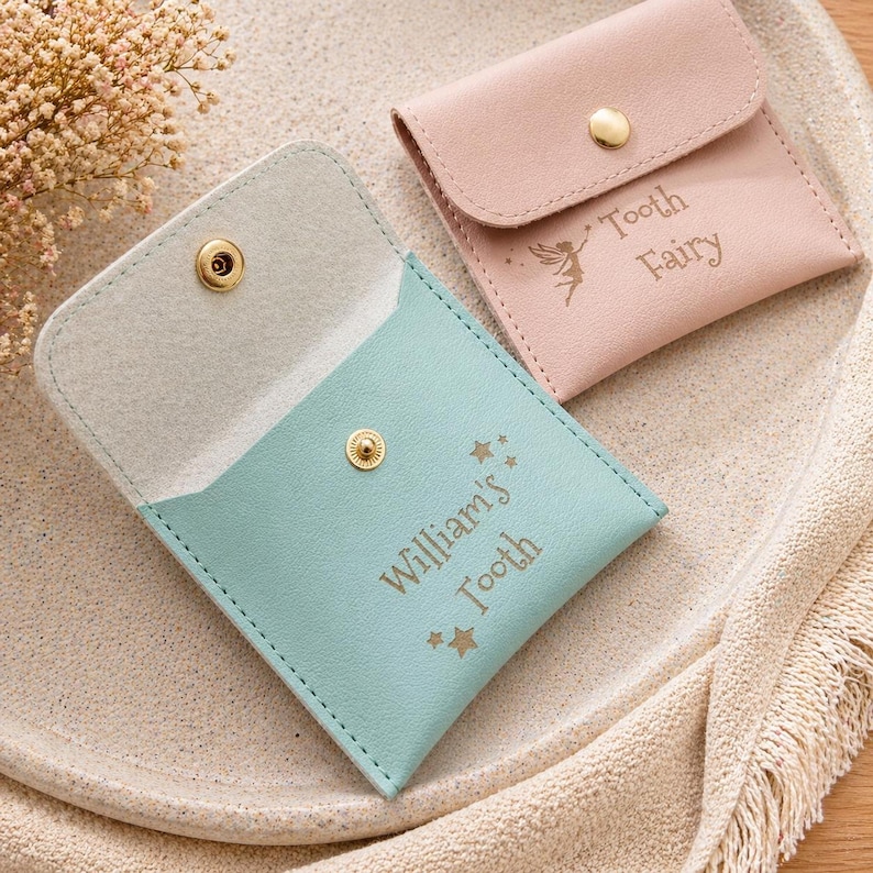 A mint green faux leather tooth fairy pouch lies open, showing its tan interior and gold text, next to a closed pink pouch on a light-colored tray.