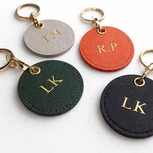 May include: Four personalised keyrings in grey, orange, green, and black. Each round leather keyring has gold-coloured initials and a gold-tone keyring. The initials are "T.M", "R.P", "L.K", and "L.K".
