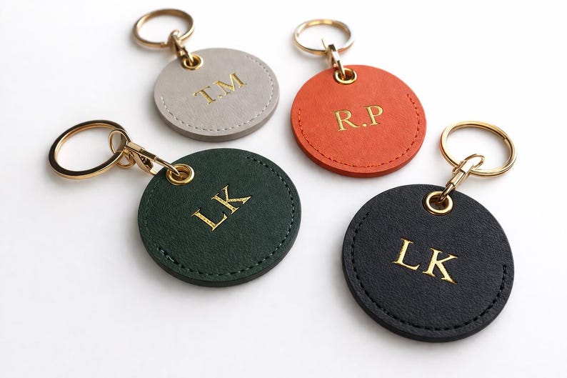 May include: Four personalised keyrings in grey, orange, green, and black. Each round leather keyring has gold-coloured initials and a gold-tone keyring. The initials are "T.M", "R.P", "L.K", and "L.K".