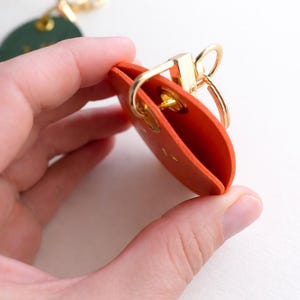 May include: An orange leather keychain holder with a gold-coloured clasp and key ring. The holder is open, revealing the interior. A green leather tag with gold hardware is visible in the background.