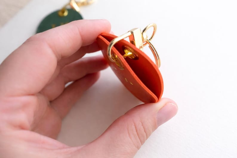May include: An orange leather keychain holder with a gold-coloured clasp and key ring. The holder is open, revealing the interior. A green leather tag with gold hardware is visible in the background.
