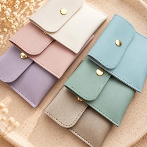 May include: A collection of six small rectangular pouches in pastel shades of white, pink, lavender, blue, green, and gray. Each pouch features a flap closure secured with a gold-toned metal snap button, arranged in two overlapping rows.