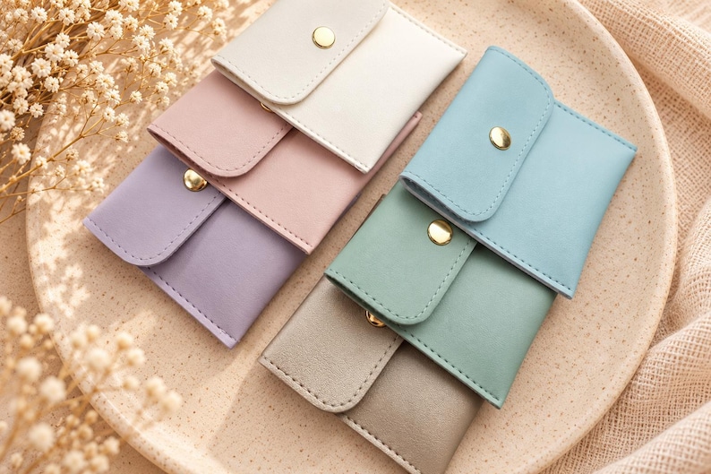 May include: A collection of six small rectangular pouches in pastel shades of white, pink, lavender, blue, green, and gray. Each pouch features a flap closure secured with a gold-toned metal snap button, arranged in two overlapping rows.