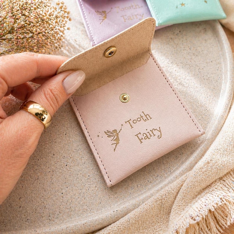A hand with a gold ring holds open a pink faux leather tooth fairy pouch, revealing its tan interior and the gold text "Tooth Fairy" with a fairy graphic.