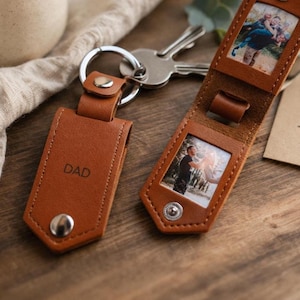 May include: A tan leather keychain with the word "DAD" embossed on it. The keychain is attached to a silver keyring and has a snap closure. Another leather piece holds two small photos. The background is a wooden surface.