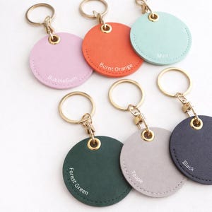 May include: A selection of circular keyrings in shades such as pink, orange, mint, forest green, taupe, and black. Each keyring features a gold-tone ring and clasp. The colour names are printed on each keyring.