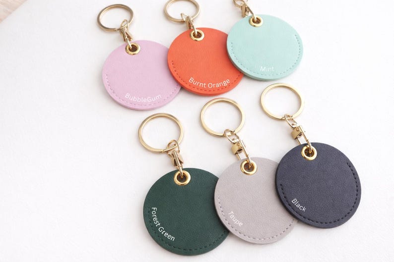 May include: A selection of circular keyrings in shades such as pink, orange, mint, forest green, taupe, and black. Each keyring features a gold-tone ring and clasp. The colour names are printed on each keyring.