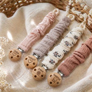 May include: Four dummy clips featuring gathered fabric straps in solid pink, solid taupe, cream with a floral print, and solid dusty rose. Each strap attaches to a round wooden clip engraved with names including Zara, Margot, and Harper.
