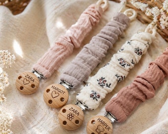 May include: Four pacifier clips featuring gathered fabric straps in solid pink, solid taupe, cream with a floral print, and solid dusty rose. Each strap attaches to a round wooden clip engraved with names including Zara, Margot, and Harper.