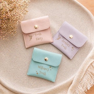 Three closed faux leather tooth fairy pouches in pink, purple, and mint green, each with a gold snap and personalized text, are arranged on a light-colored tray.