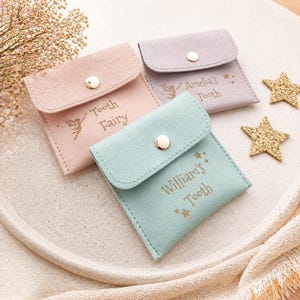 Three closed faux leather tooth fairy pouches in pink, purple, and mint green, each with a gold snap and personalized text, are arranged on a light-colored tray with two gold star decorations.