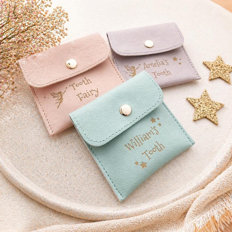 Three closed faux leather tooth fairy pouches in pink, purple, and mint green, each with a gold snap and personalized text, are arranged on a light-colored tray with two gold star decorations.