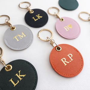 May include: A selection of personalised keyrings in various colours, including black, grey, pink, and orange. Each keyring features gold-coloured initials or a heart symbol. The keyrings have gold-coloured key rings.