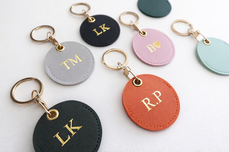 May include: A selection of personalised keyrings in various colours, including black, grey, pink, and orange. Each keyring features gold-coloured initials or a heart symbol. The keyrings have gold-coloured key rings.