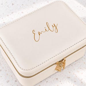 Könnte beinhalten: Eine rechteckige, cremefarbene Schmuckschatulle mit glatter Oberfläche und goldenem Reißverschluss. Auf der Oberseite ist der Name Emily in goldener Schreibschrift zu sehen. Das Etui weist sichtbare Nähte an den Kanten auf.