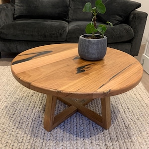 May include: A circular coffee table made of light-coloured wood, with a dark stain detail. The table has a cross-shaped base and is approximately 76 cm in diameter. A small potted plant sits on the table's surface.