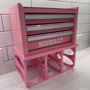 May include: A pink metal tool chest with three drawers featuring silver handles and the white text KOBALT printed on the bottom drawer. The chest is supported by a pink, open-frame, rectangular stand with oval cutouts on the sides.