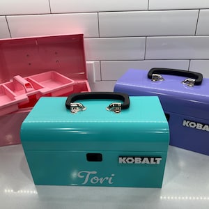 May include: Three colorful metal toolboxes in pink, teal, and purple. The teal toolbox has the name "Tori" and the brand name "KOBALT" on it. The pink toolbox is open, revealing interior compartments. Each toolbox has a black handle.