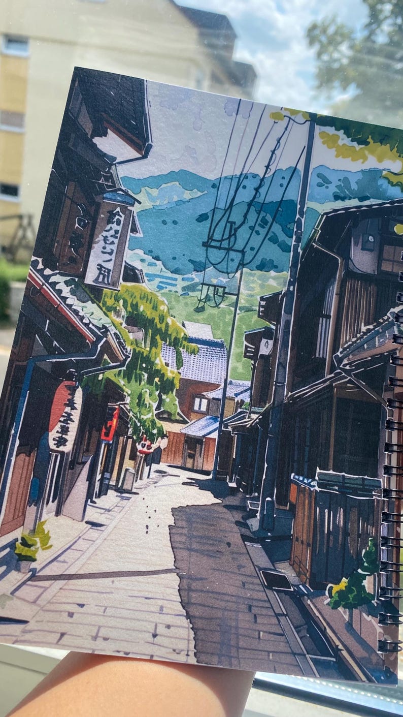 May include: A spiral-bound sketchbook page featuring a detailed marker illustration of a narrow Japanese street. The scene depicts traditional wooden buildings, hanging red lanterns, vertical signage with Japanese text, and power lines.