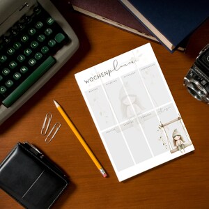 May include: A weekly planner with the German text "Wochenplaner" is displayed on a wooden desk. The planner features illustrations of a girl on a swing and other decorative elements. A vintage typewriter, pencil, and camera are also on the desk.
