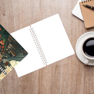 May include: An open spiral notebook with blank white pages next to a closed notebook with a colorful illustration. A cup of coffee, a pen, and glasses are on a wooden surface.