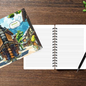 May include: A spiral-bound notebook with a white interior featuring horizontal lines. The cover displays a colorful illustration of a traditional street scene with wooden buildings, green trees, and a blue sky. The word Notebook is printed on the cover.