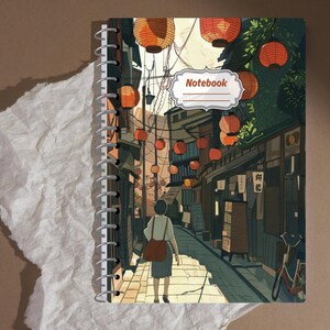 May include: Spiral-bound notebook featuring an illustrated Japanese street scene with hanging lanterns. The cover art depicts a person walking down a cobblestone street lined with shops. The word "Notebook" is in a white label on the cover.