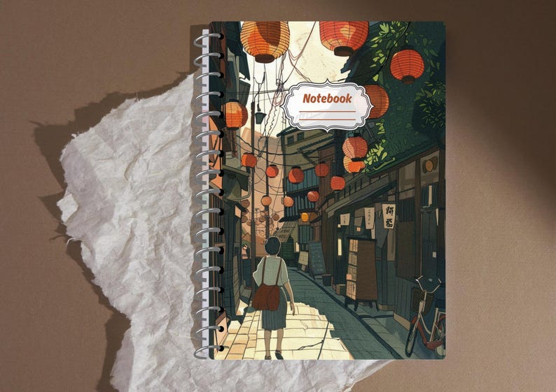 May include: Spiral-bound notebook featuring an illustrated Japanese street scene with hanging lanterns. The cover art depicts a person walking down a cobblestone street lined with shops. The word "Notebook" is in a white label on the cover.