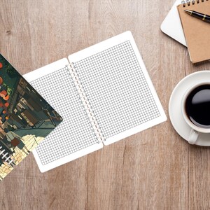 May include: An open notebook with grid paper sits next to a closed notebook with a colorful illustration. A cup of coffee, glasses, a pen, and a laptop are on a wooden surface. The closed notebook has the word "Notebook" on it.