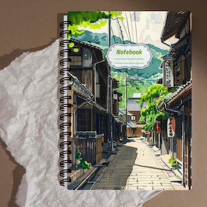 May include: A spiral-bound notebook featuring an illustration of a narrow Japanese street with traditional wooden buildings, green trees, and power lines. The cover includes a white label with the word Notebook printed in green text with lines below.
