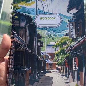 May include: A spiral-bound notebook featuring a colorful illustration of a narrow Japanese street with traditional wooden buildings, green trees, and mountains in the background. The cover includes a white label with the word Notebook printed on it.