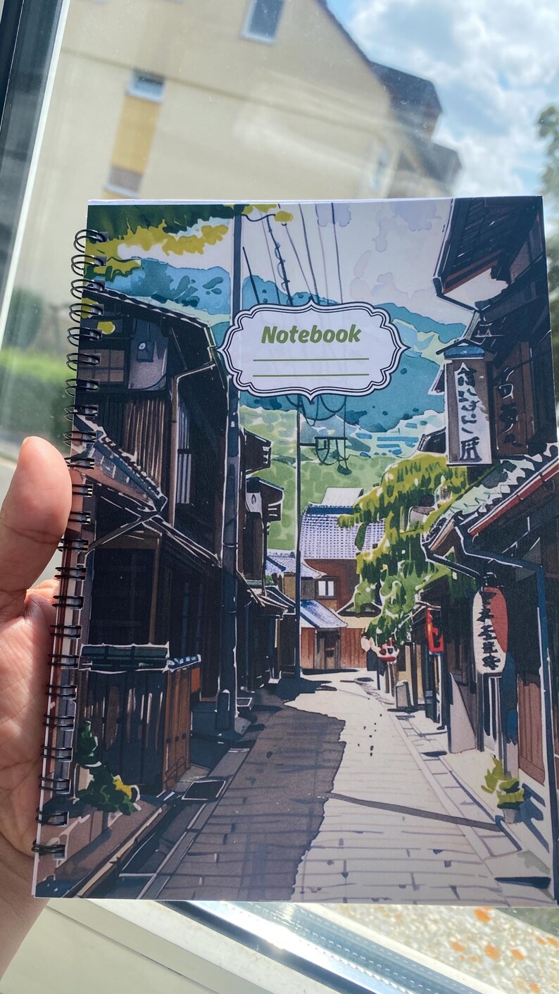 May include: A spiral-bound notebook featuring a colorful illustration of a narrow Japanese street with traditional wooden buildings, green trees, and mountains in the background. The cover includes a white label with the word Notebook printed on it.