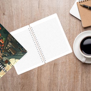 May include: An open spiral notebook with dotted pages sits next to a closed notebook with a colorful illustration. A cup of coffee, glasses, a pen, and a laptop are on a wooden surface. The closed notebook has the word "Notebook" on it.