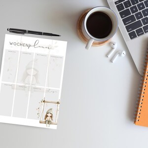 May include: A weekly planner with the German text "Wochenplaner" and illustrations of a girl on a swing and a girl in a hooded outfit. A black pen, a cup of coffee, a laptop, a spiral notebook, and a pencil are also on the white surface.