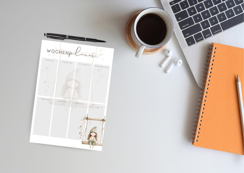 May include: A weekly planner with the German text "Wochenplaner" and illustrations of a girl on a swing and a girl in a hooded outfit. A black pen, a cup of coffee, a laptop, a spiral notebook, and a pencil are also on the white surface.