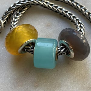 May include: A silver chain bracelet with three colorful beads. The beads are yellow, turquoise, and gray, and have a faceted or cylindrical shape. The beads are strung on a silver chain. The brand name "BEADACKER" is in the top left corner.