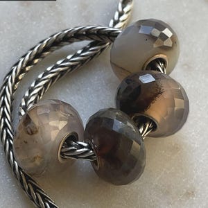 Op de afbeelding: Close-up van een zilveren ketting met facetgeslepen kralen in neutrale tinten. De kralen zijn in verschillende bruin- en grijstinten en hebben een geometrische snit. Ze zijn geregen op een zilveren ketting.