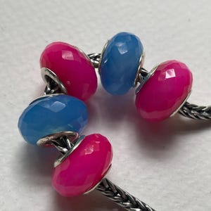 May include: A silver chain bracelet featuring five faceted, round beads in alternating hot pink and light blue colors. The beads are arranged along the metallic chain, each showing a polished, multi-faceted surface that reflects light.