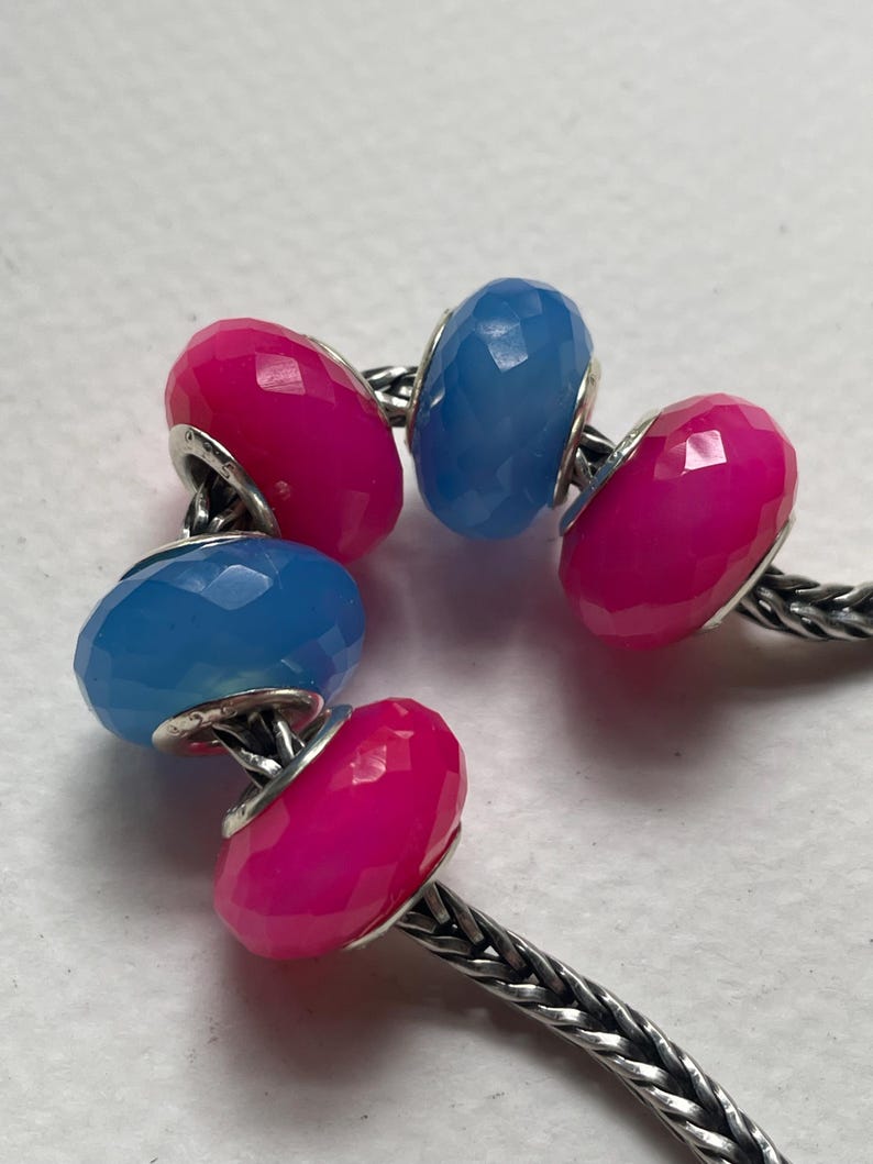 May include: A silver chain bracelet featuring five faceted, round beads in alternating hot pink and light blue colors. The beads are arranged along the metallic chain, each showing a polished, multi-faceted surface that reflects light.