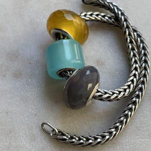Op de afbeelding: Een zilveren armband met drie kleurrijke kralen: geel, turquoise en grijs. De armband heeft een gevlochten kettingontwerp en een sluiting. De merknaam "BEADACKER" staat linksboven.