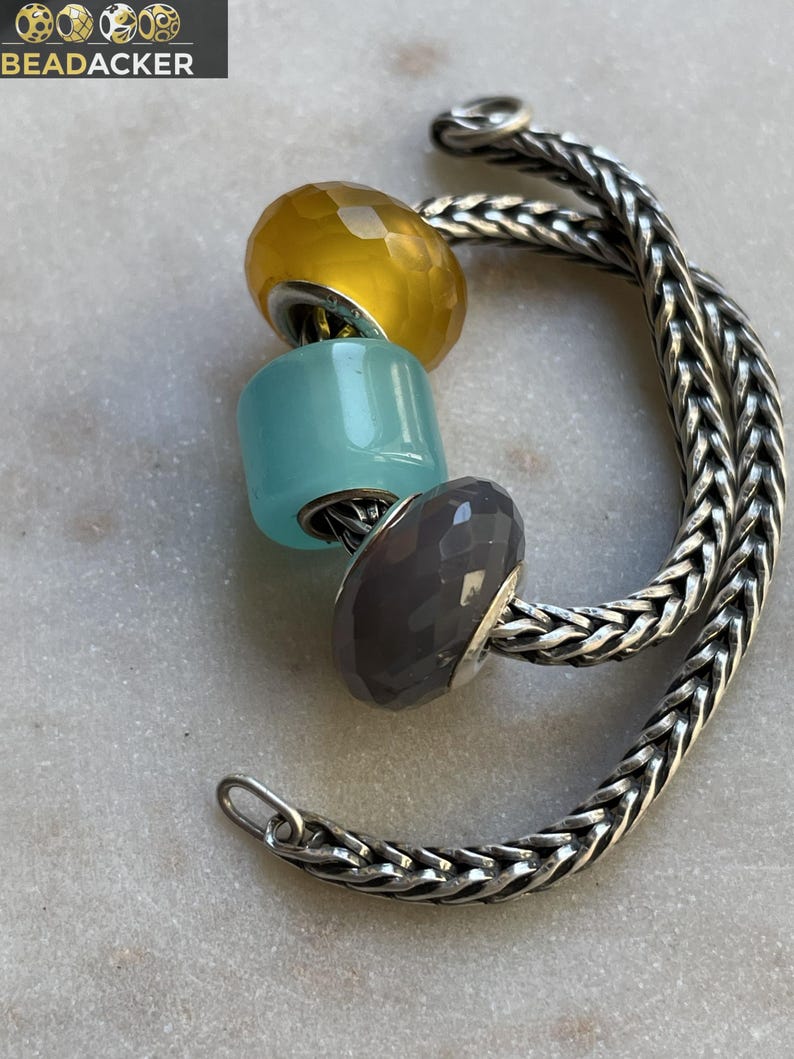 Op de afbeelding: Een zilveren armband met drie kleurrijke kralen: geel, turquoise en grijs. De armband heeft een gevlochten kettingontwerp en een sluiting. De merknaam "BEADACKER" staat linksboven.