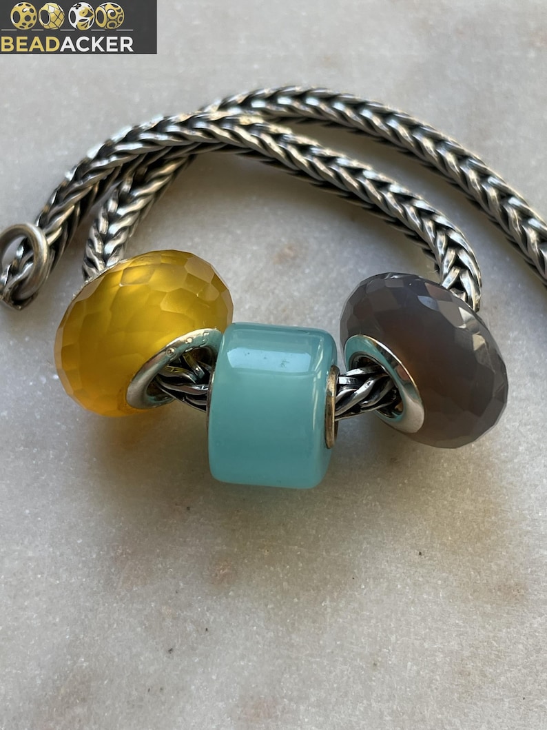 May include: A silver chain bracelet with three colorful beads. The beads are yellow, turquoise, and gray, and have a faceted or cylindrical shape. The beads are strung on a silver chain. The brand name "BEADACKER" is in the top left corner.
