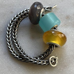 Op de afbeelding: Een zilveren kettingarmband met drie decoratieve kralen. De kralen zijn grijs, lichtblauw en geel. De armband heeft een sluiting. De merknaam "BEADACKER" staat in de linkerbovenhoek.