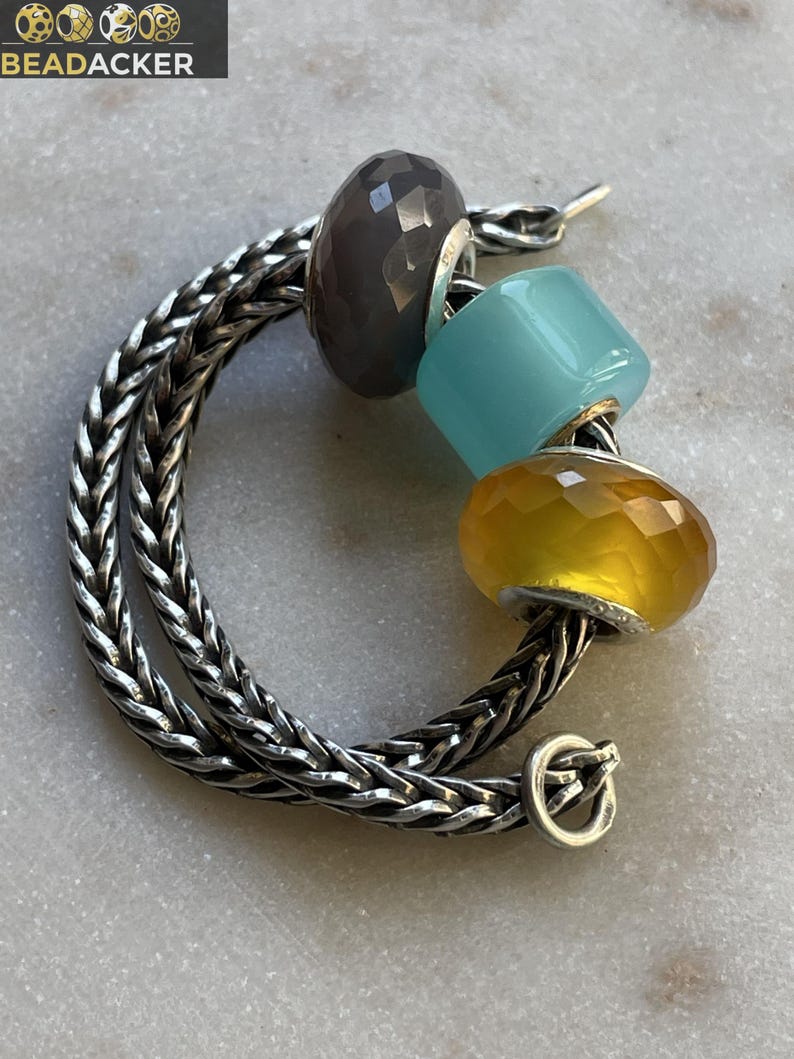 Op de afbeelding: Een zilveren kettingarmband met drie decoratieve kralen. De kralen zijn grijs, lichtblauw en geel. De armband heeft een sluiting. De merknaam "BEADACKER" staat in de linkerbovenhoek.