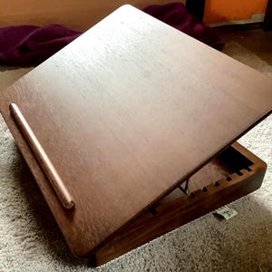 May include: A wooden adjustable laptop desk with a warm brown finish. The desk features a sloped surface and a small ledge to hold items in place. The base has a storage compartment and is designed to be sturdy.