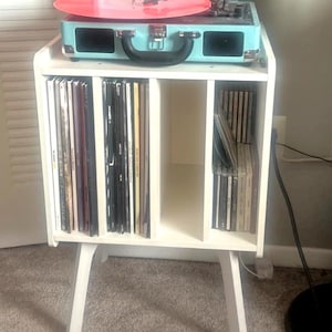 May include: A white wooden record player stand with a turquoise record player on top. A red vinyl record is on the turntable. The stand has compartments for vinyl records and CDs. The stand is on a carpeted floor.