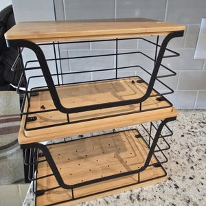 May include: A two-tiered kitchen organizer with a bamboo wood top and base. The organizer has a black metal wire frame. The top shelf is rectangular, and the bottom two shelves are square. The organizer is designed to hold kitchen items.