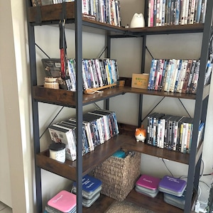 May include: A dark brown and black metal corner bookshelf filled with DVDs, decorative items, and storage containers. The shelves are made of dark wood and the frame is black metal. A woven basket and several colorful containers are visible.