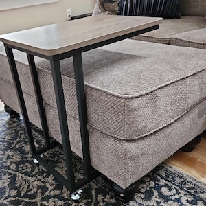 May include: A side table with a light wood-tone top and a black metal frame. The table is designed to slide under a sofa or chair, and it has small wheels for easy movement. The table's dimensions are approximately 24 inches long, 12 inches wide, and 24 inches high.