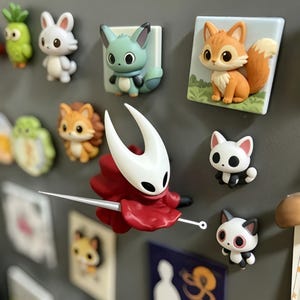 Puede incluir: Una colección de imanes de personajes sobre una superficie gris, destacando una figura con máscara blanca y capa roja que sostiene una aguja, junto a imanes de animales más pequeños como un zorro, gatos y un conejo.